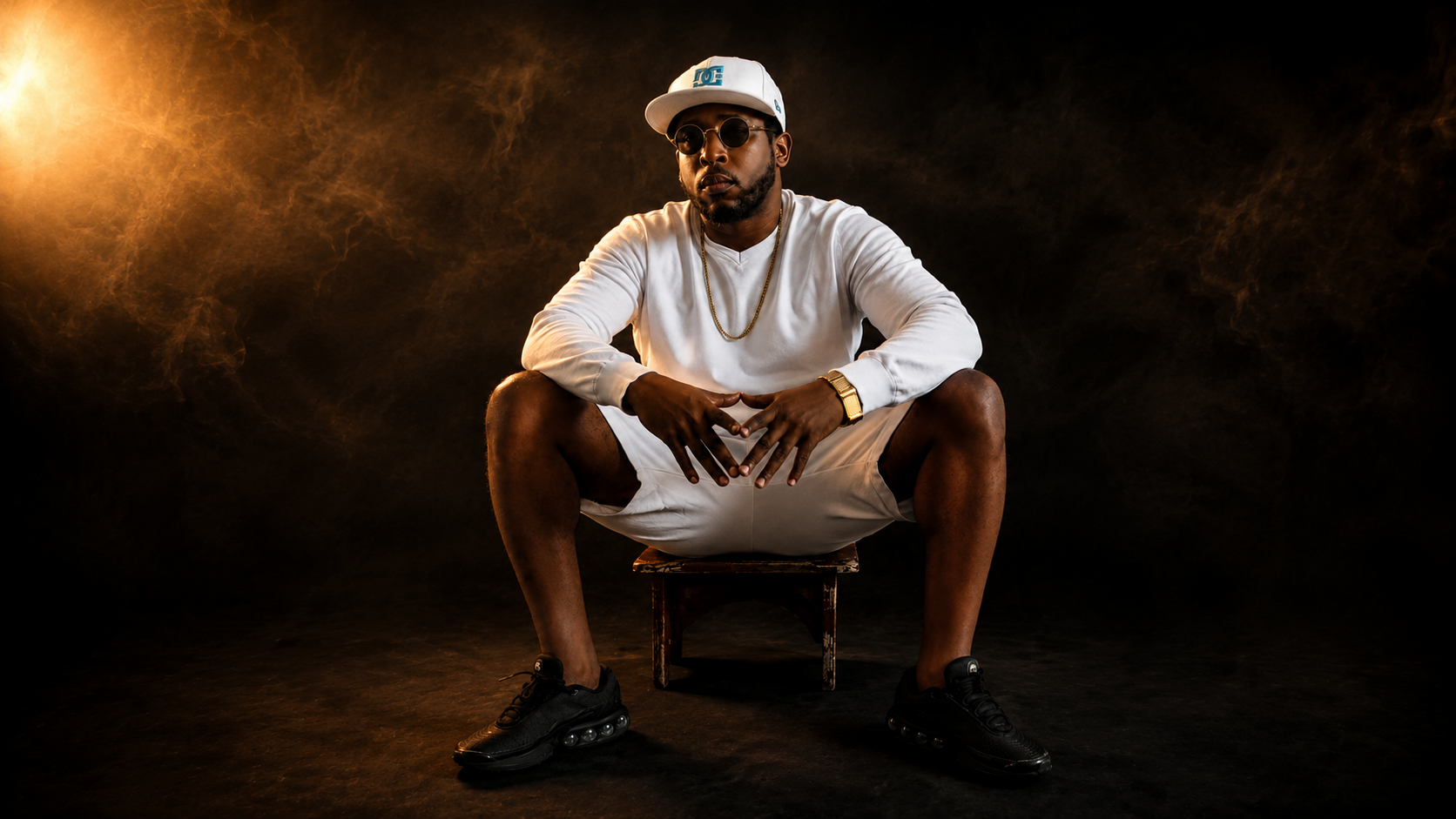 l'artiste Ochomane habillé d'une casquette blanche un sweat à manche longue blanc et un short blanc, assis sur un petit banc