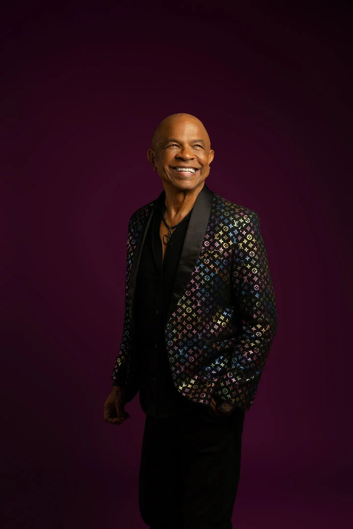 Francky Vincent smiling in a jacket with colourful motifs against a purple background.