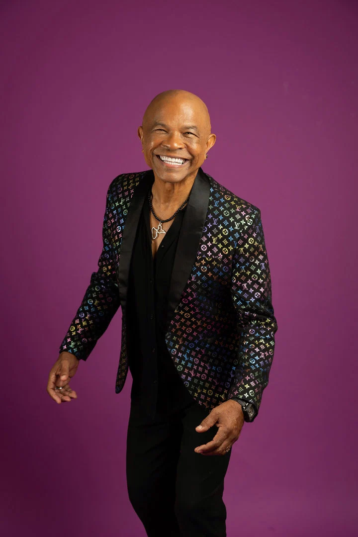 Francky Vincent smiling at the camera, wearing a black shirt and patterned jacket against a purple background.